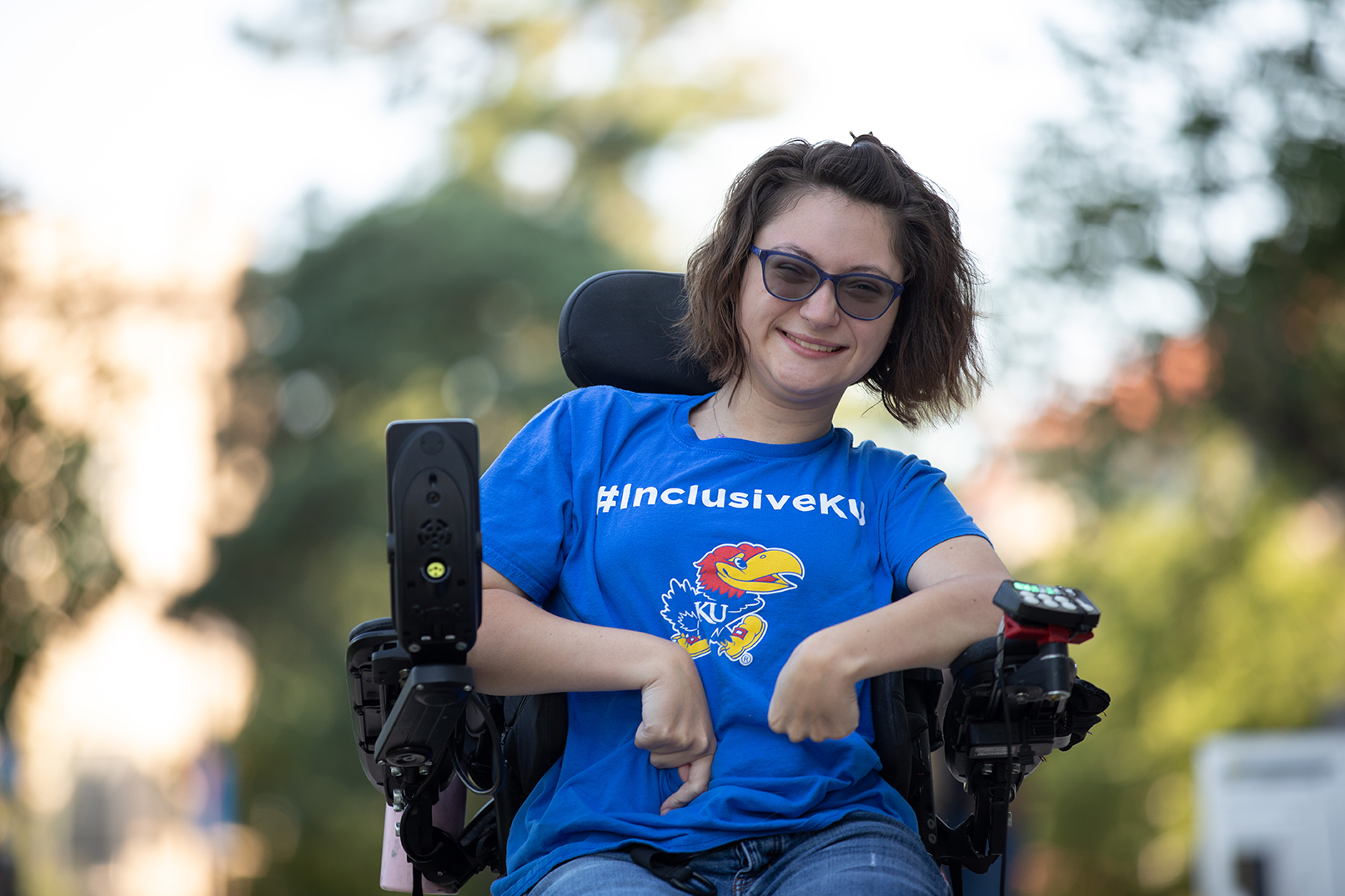 KU student Carleigh Lavoy sits smiling in her wheelchair with a shirt that reads "InclusiveKU